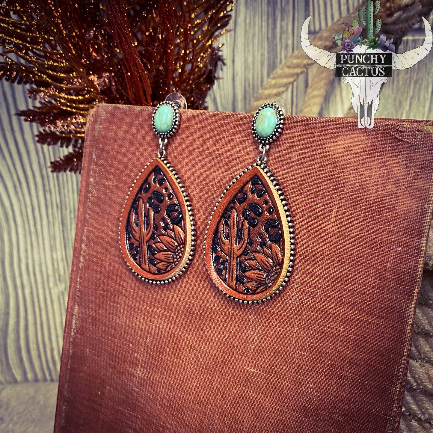 Western leather tooled earrings. Tooled leather is tear drop shaped. Tooled into the leather is a cactus and a sunflower, with a cheetah print back ground.
